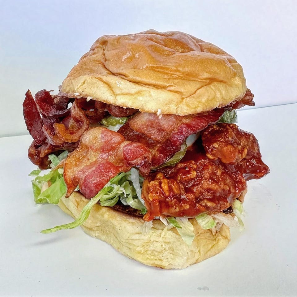 A Jojo sandwich with crispy bacon, saucy chicken, lettuce, and pickles on a brioche bun.