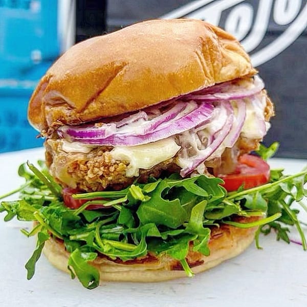 A Jojo sandwich with fried chicken, red onions, cheese, tomatoes, and arugula on a bun.