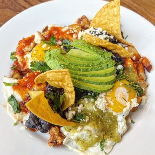 Huevos rancheros with fried eggs, avocado, black beans, salsa, and tortilla chips on a white plate.